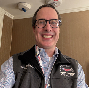 A smiling man with glasses, wearing a blue checkered shirt and a black vest, in a room with beige walls.