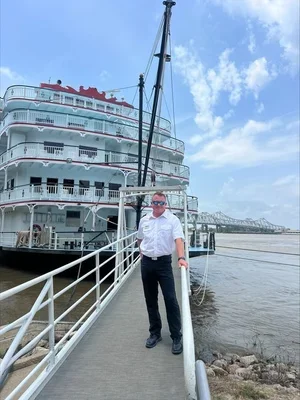 A man in a white shirt and sunglasses standing on a dock next to a large cruise ship with multiple decks, under a partly cloudy sky.