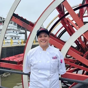 A woman in a white chef's coat and black cap standing in front of a red and white Ferris wheel at a waterfront.