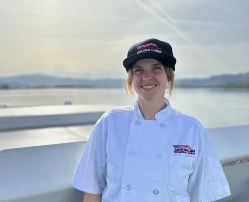 Woman in a white chef's coat and black cap smiling near water with a city skyline in the background.