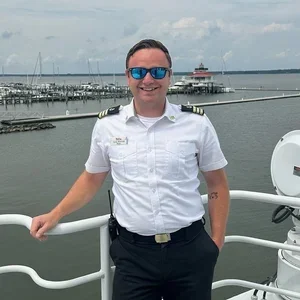A man dressed as a captain or crew member standing on a boat, smiling, with a marina and a lighthouse in the background.