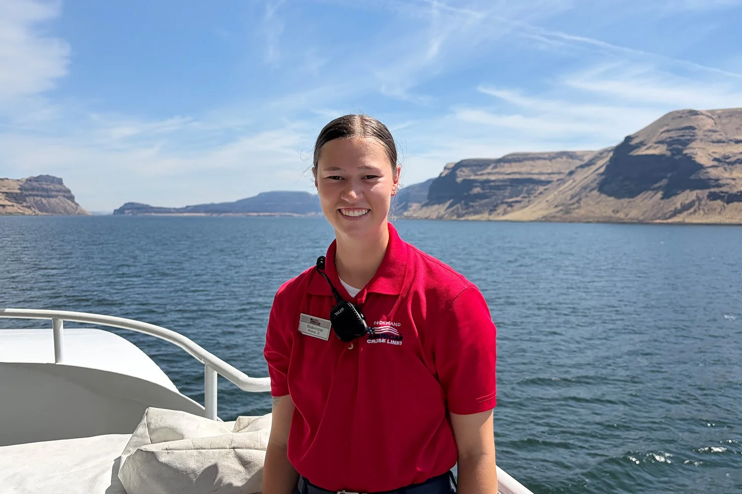 Deckhand aboard an American Cruise Lines ship standing on the deck with scenic cliffs and water in the background.