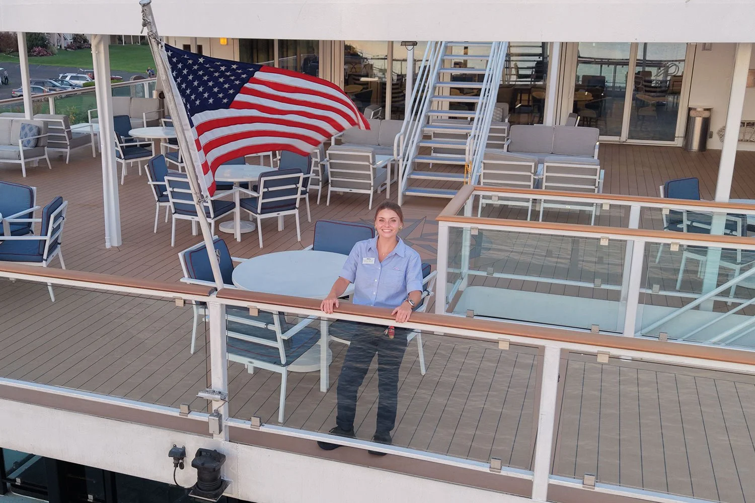 Hospitality crew member standing on the deck of an American Cruise Lines ship beside an American flag and outdoor seating area.