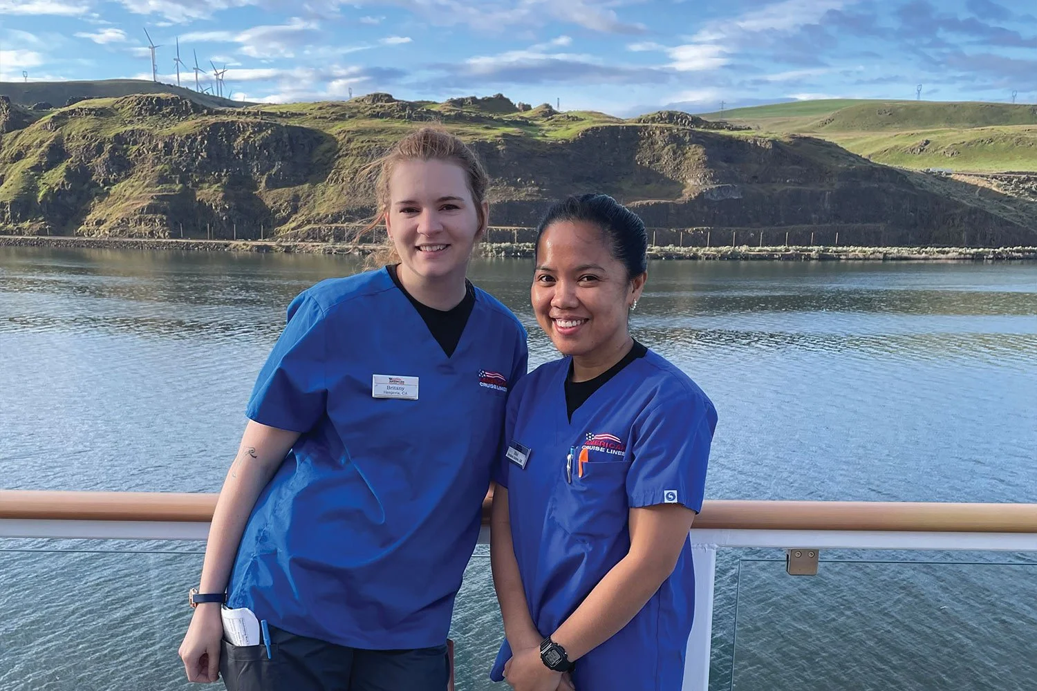 Two women in blue nursing scrubs standing on a boat deck with a body of water and green hills with wind turbines in the background.