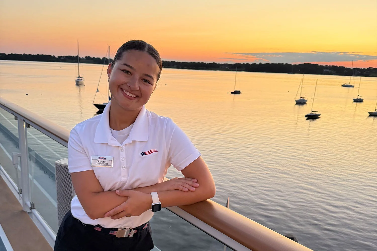 A young woman with dark hair tied back, smiling, wearing a white polo shirt with a name tag that says Shyann and a small American flag logo, stands on the deck of a boat at sunset with a calm body of water and sailboats in the background.