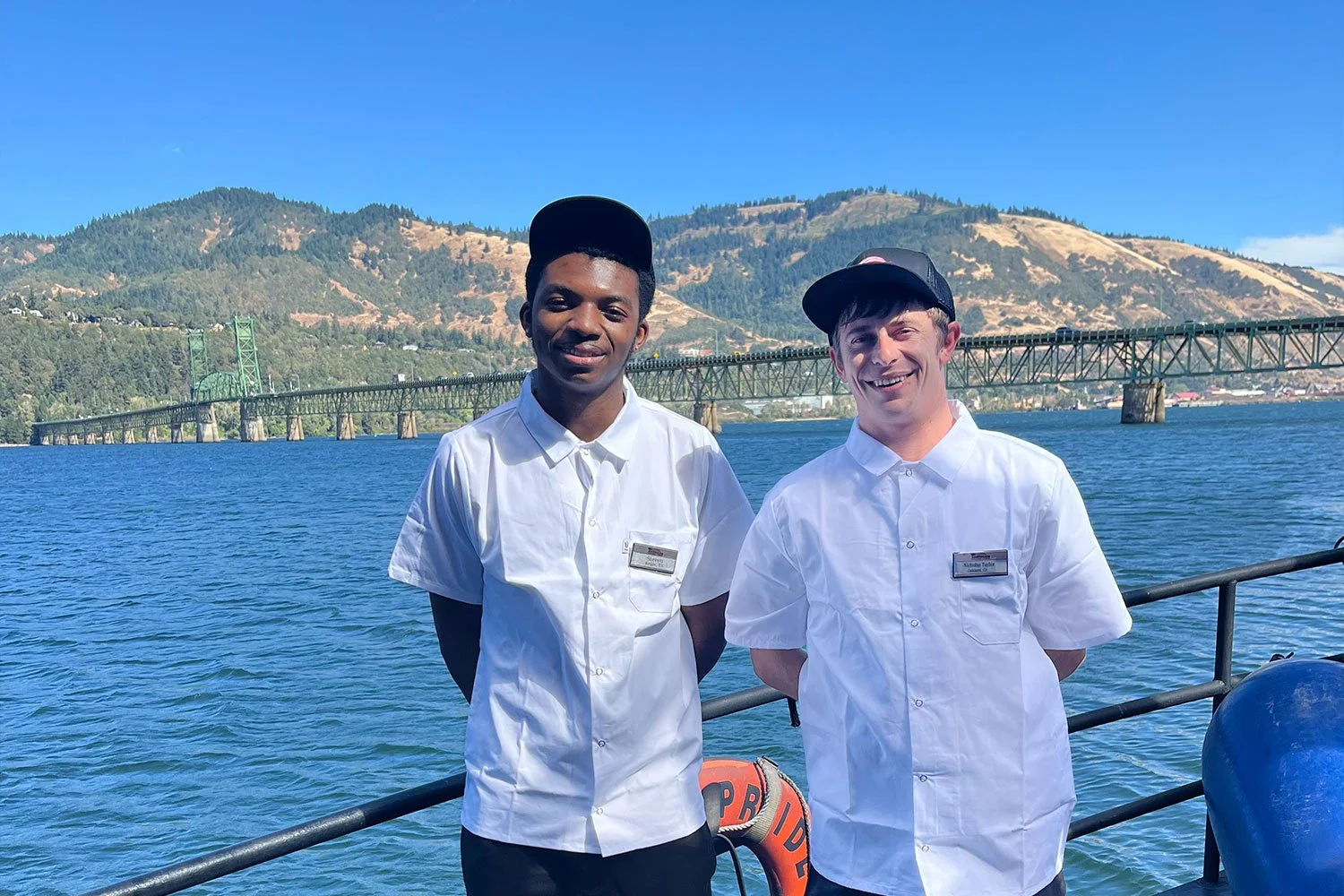 Two smiling galley stewards in white uniforms and black caps stand on a ship deck with a large bridge, water, and mountains behind them.
