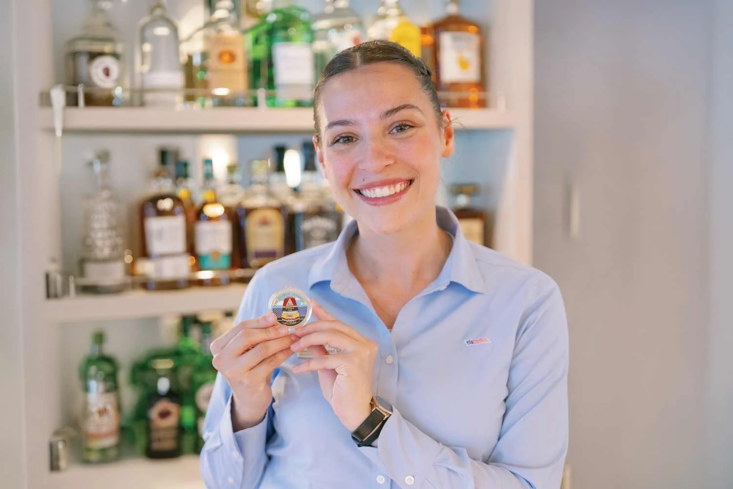A smiling hotel general manager in a light blue button-down shirt holding up a small round emblem, standing in front of a well-stocked lounge bar display.