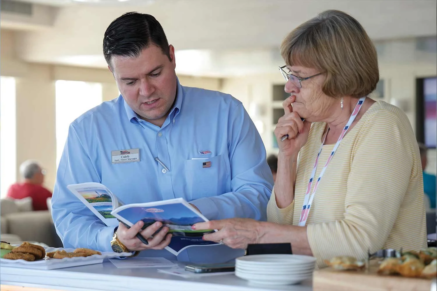 An assistant hotel manager in a light blue collared shirt reviews a cruise brochure alongside a female passenger indoors.