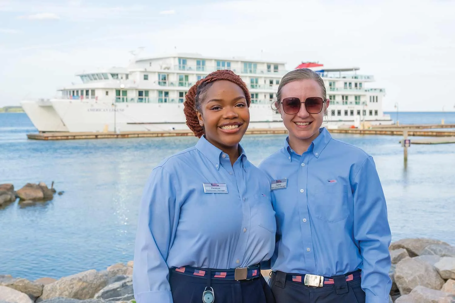Two smiling American Cruise Lines managers in light blue collared shirts stand outdoors by the water with a cruise ship docked in the background.