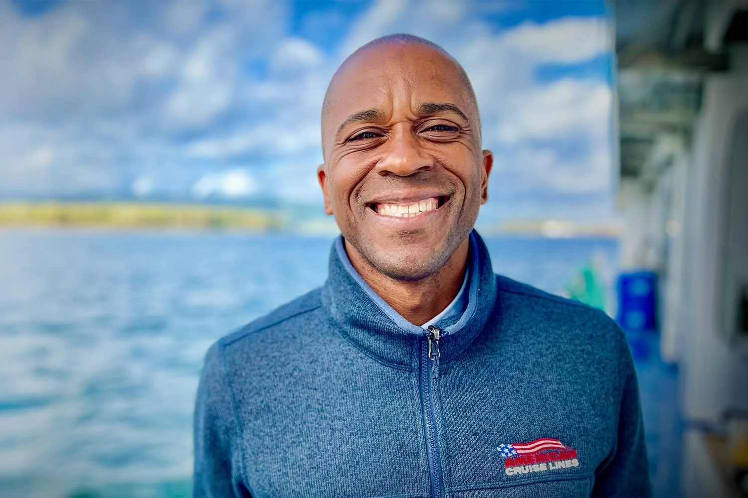 A smiling man on a boat with water and cloudy sky in the background, wearing a blue zip-up jacket with a logo that says 'AMERICAN CRUISE LINES'.