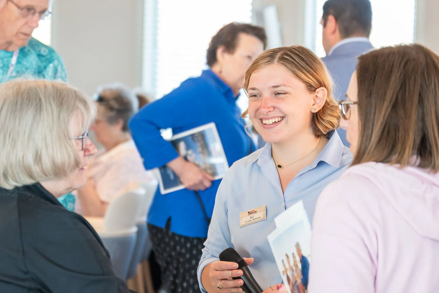 A woman with short red hair smiling while holding a microphone, engaging in conversation with a woman with brown hair wearing glasses. Other people are interacting and talking in the background at what appears to be a social or professional event.