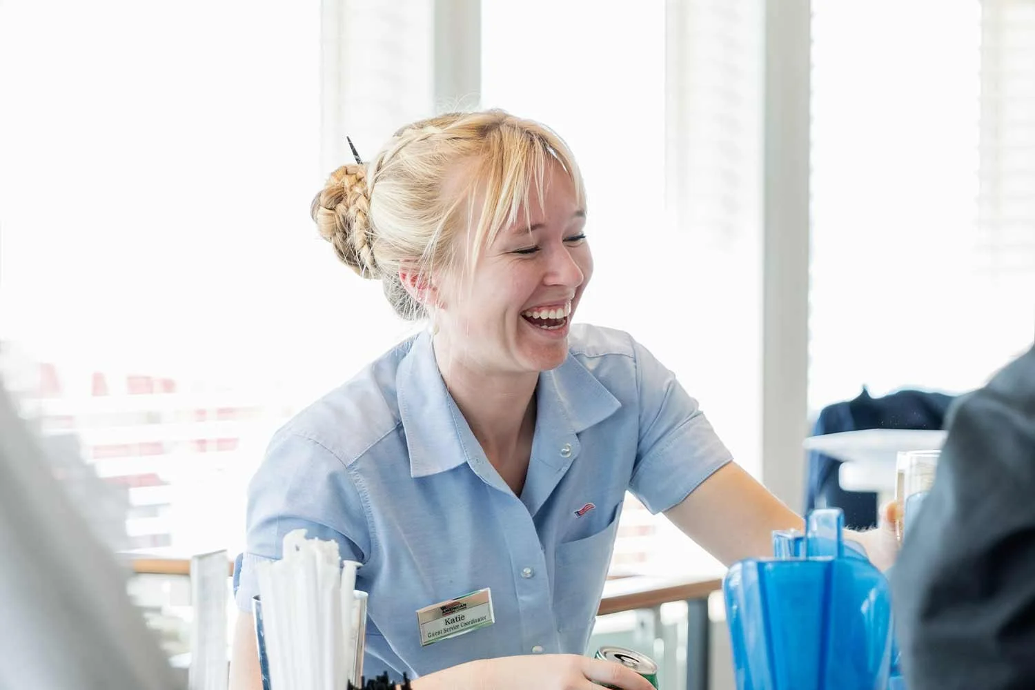 A woman with blonde hair in a braided updo, smiling and wearing a light blue shirt, sitting at a table with a name tag that reads 'Katie.' She appears to be working as a server in a bright, cheerful environment.