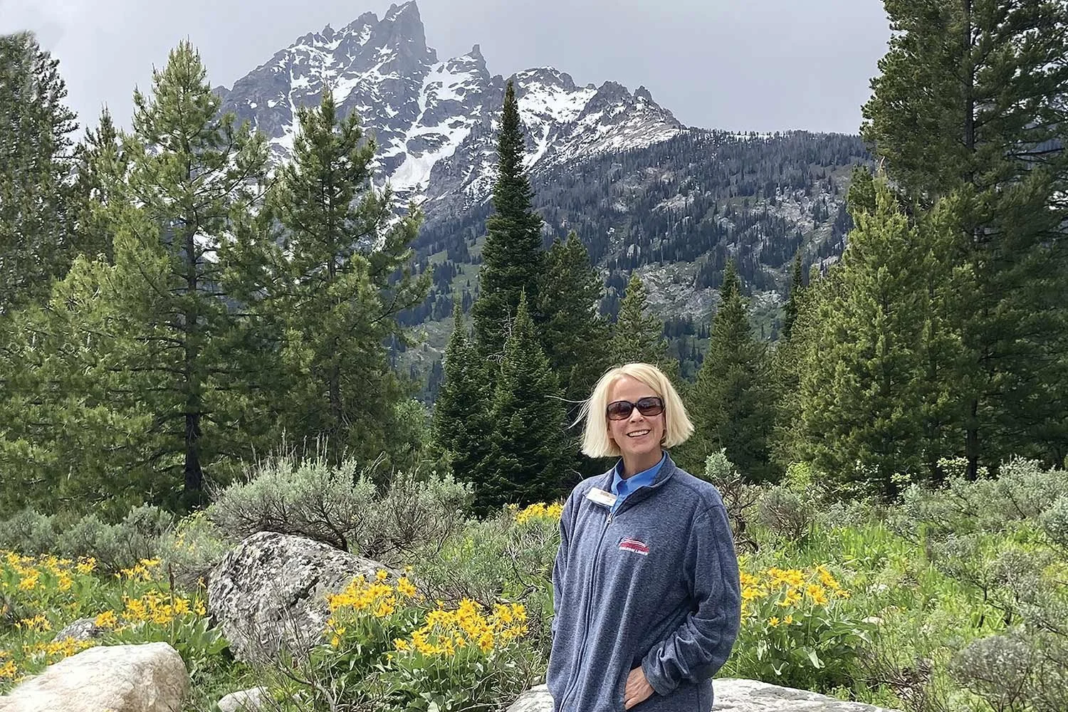 A smiling woman with blonde hair and sunglasses standing in a lush green mountain landscape with snow-capped peaks in the background, surrounded by trees and yellow flowers.