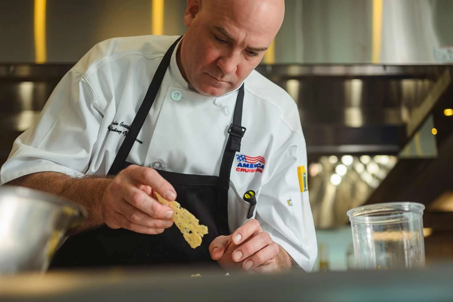 An executive chef wearing a white chef coat and a black American Cruise Lines apron carefully plates a dish in a ship galley.