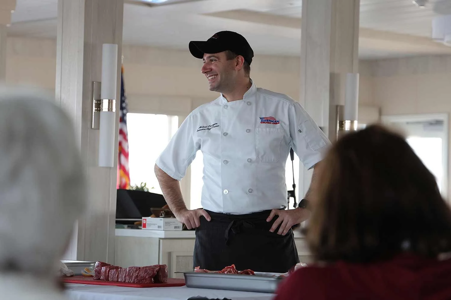 An executive sous chef in a white chef coat and black cap smiles while leading a culinary demonstration for passengers.