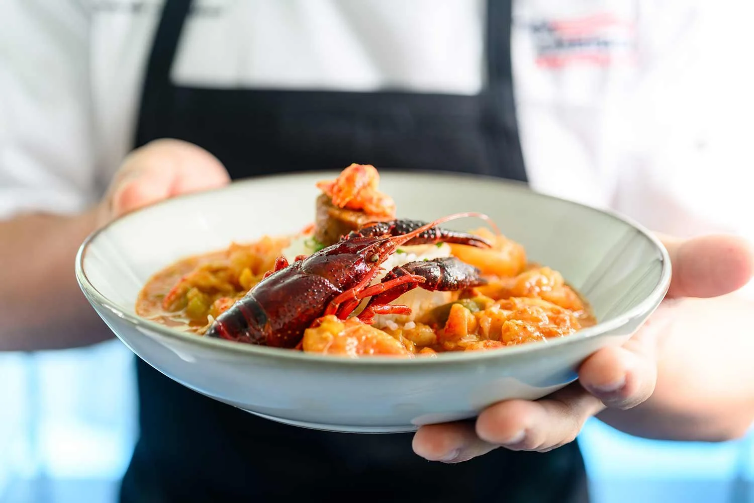 A chef holds out a freshly prepared dish featuring whole crawfish, seafood, and rice served in a shallow bowl.