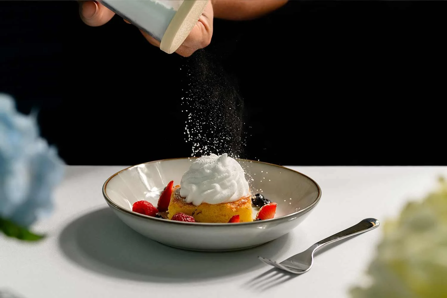 A person sprinkling powdered sugar on a dessert topped with whipped cream and surrounded by fresh berries in a white bowl, with a spoon on the side.