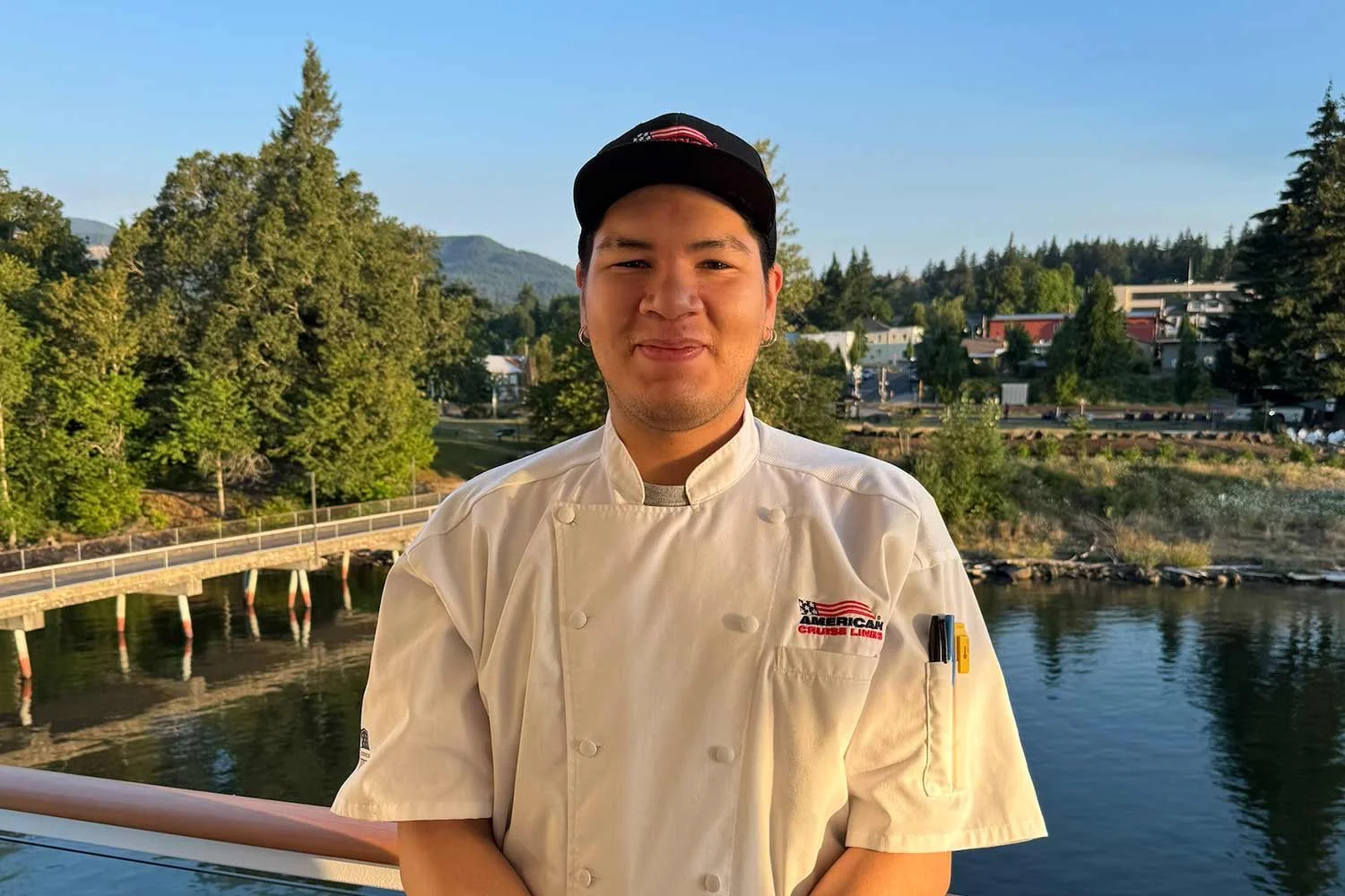 A young man in a chef's uniform standing outdoors with a river, trees, and mountains in the background.