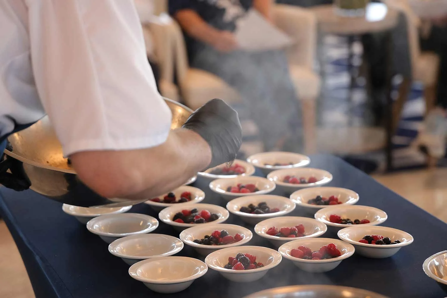 A chef preparing small bowls of dessert topped with mixed berries at a table in a restaurant setting.