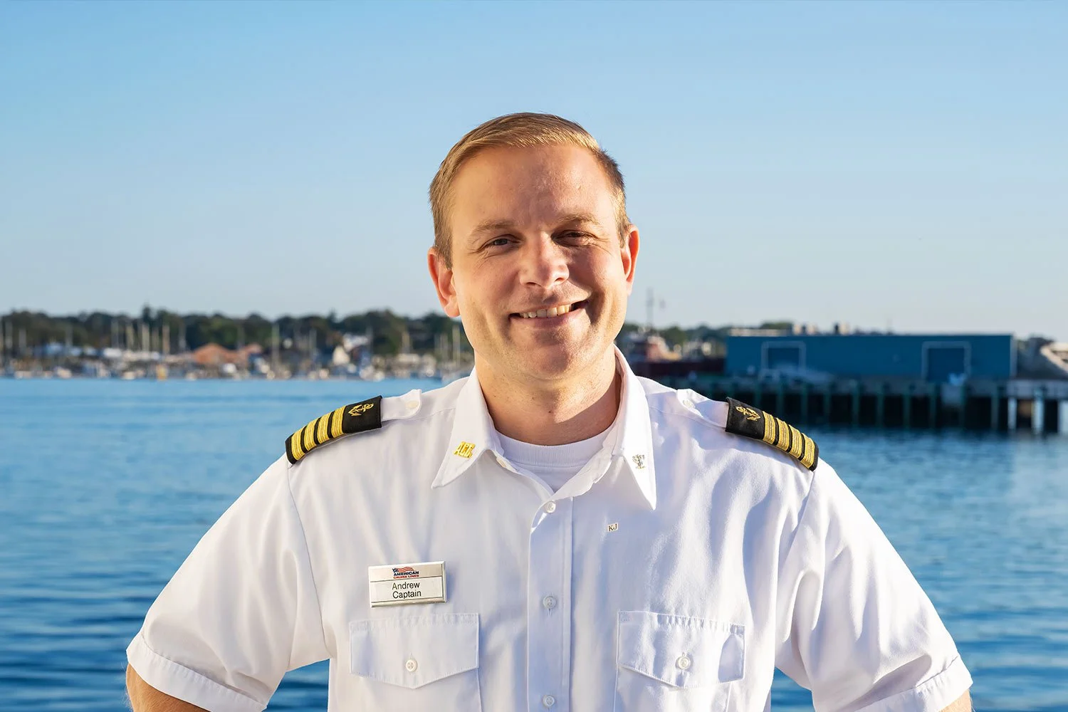 A smiling ship captain wearing a white uniform short-sleeve shirt with shoulder epaulets stands outdoors with blue water and a harbor in the background.