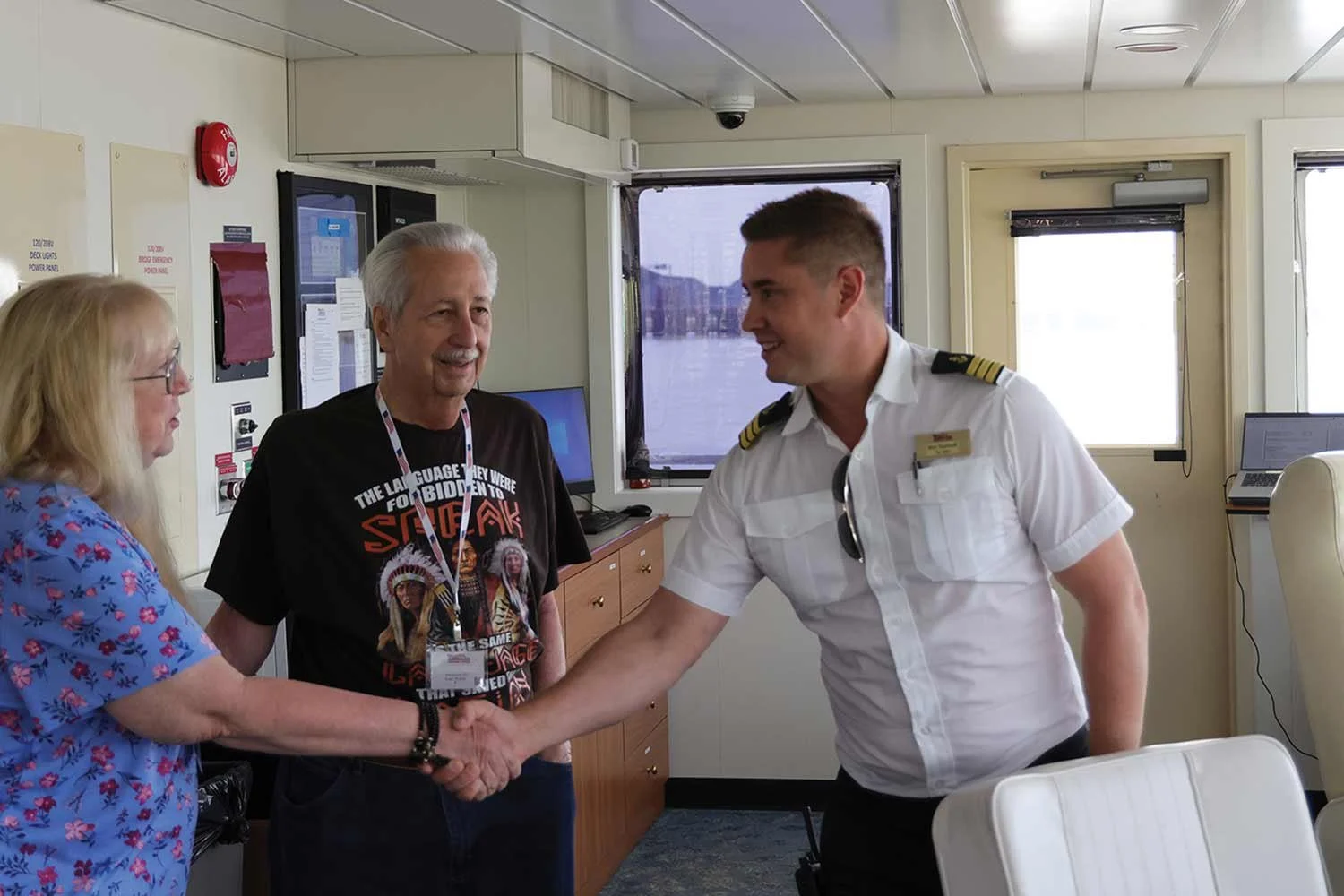 A smiling ship captain wearing a white uniform short-sleeve shirt with shoulder epaulets stands outdoors with blue water and a harbor in the background.