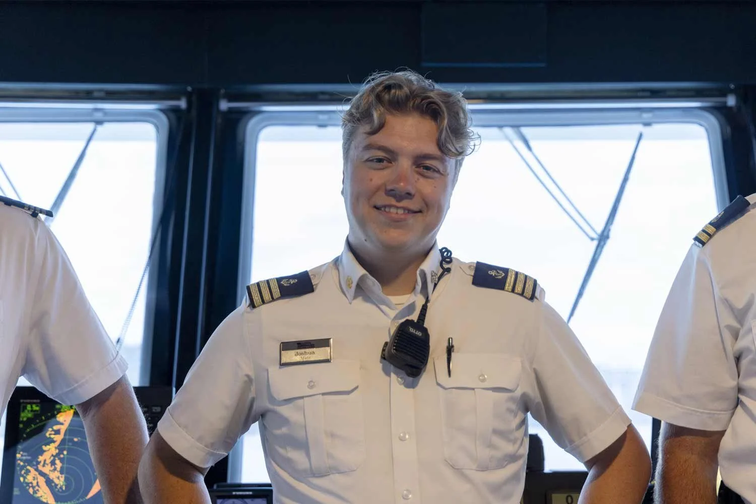 A smiling ship mate wearing a white short-sleeve uniform shirt with shoulder epaulets and a radio microphone stands inside the ship's bridge.