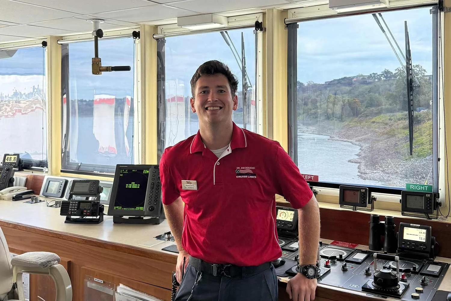 A young man in a red shirt standing inside a ship's bridge with nautical equipment and large windows showing a shoreline and water outside.