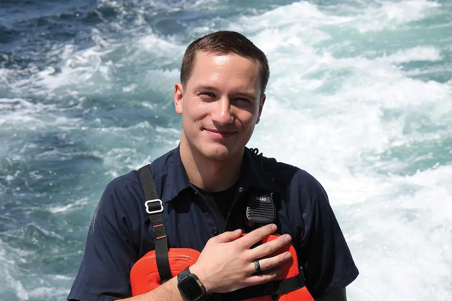 A smiling ship engineer in a dark work shirt and orange life vest stands on deck with churning blue water in the background.