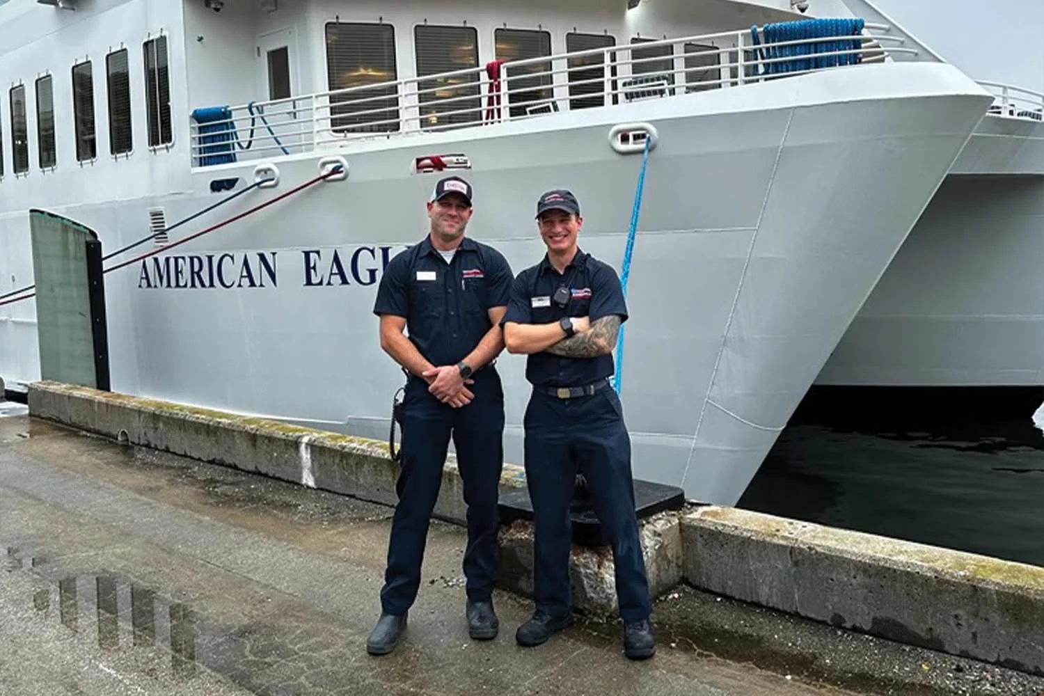 Two smiling ship engineers wearing navy work uniforms and caps stand side-by-side on a dock in front of the American Eagle cruise ship.
