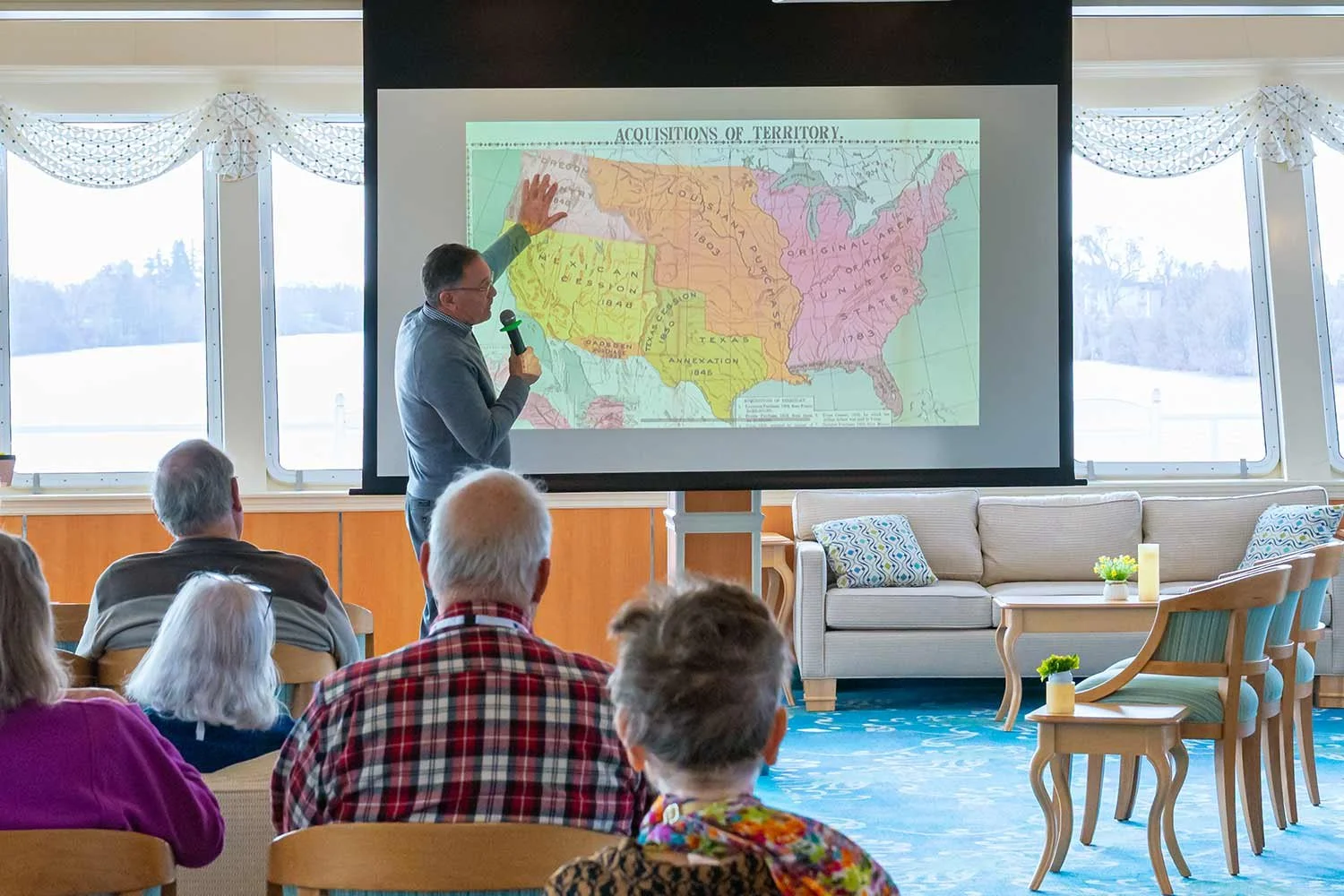 A speaker holds a microphone and points to a map projection of U.S. territorial acquisitions during a presentation to seated cruise passengers in a ship lounge.