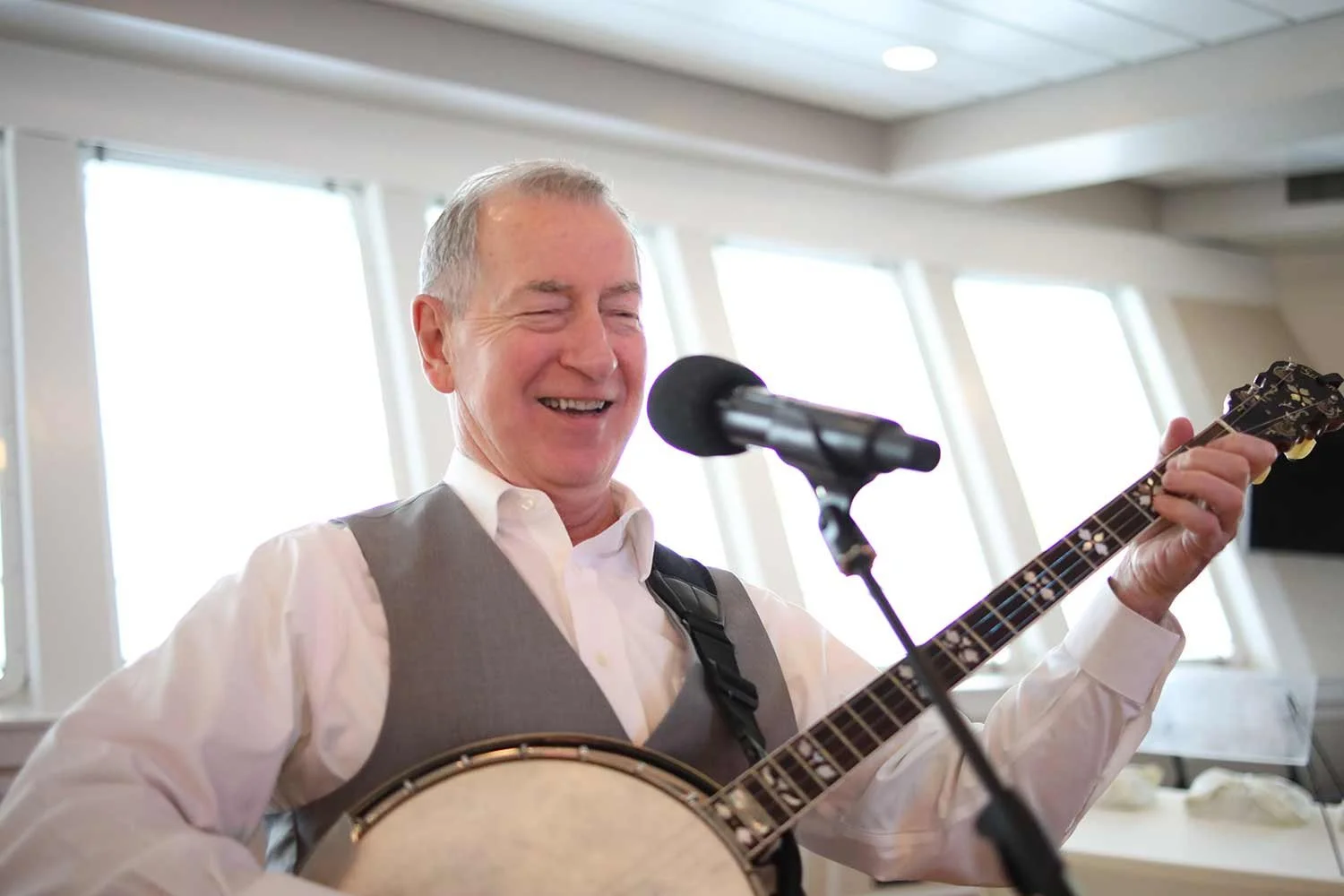 A smiling resident entertainer in a vest and dress shirt plays a banjo and sings into a microphone.