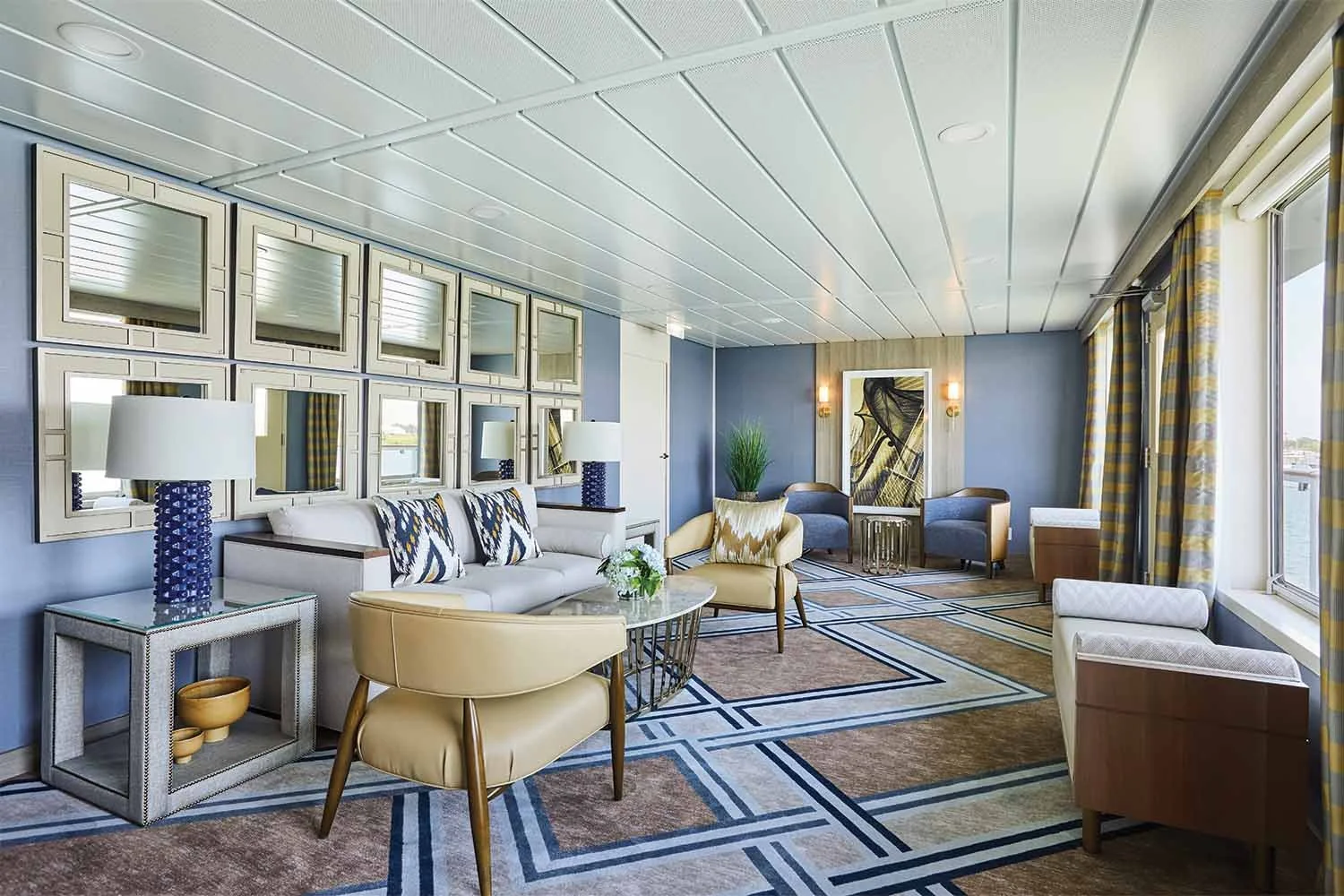 Bright hotel lounge with colorful furniture, framed mirrors on the wall, large windows with yellow and gray curtains, and contemporary decor.
