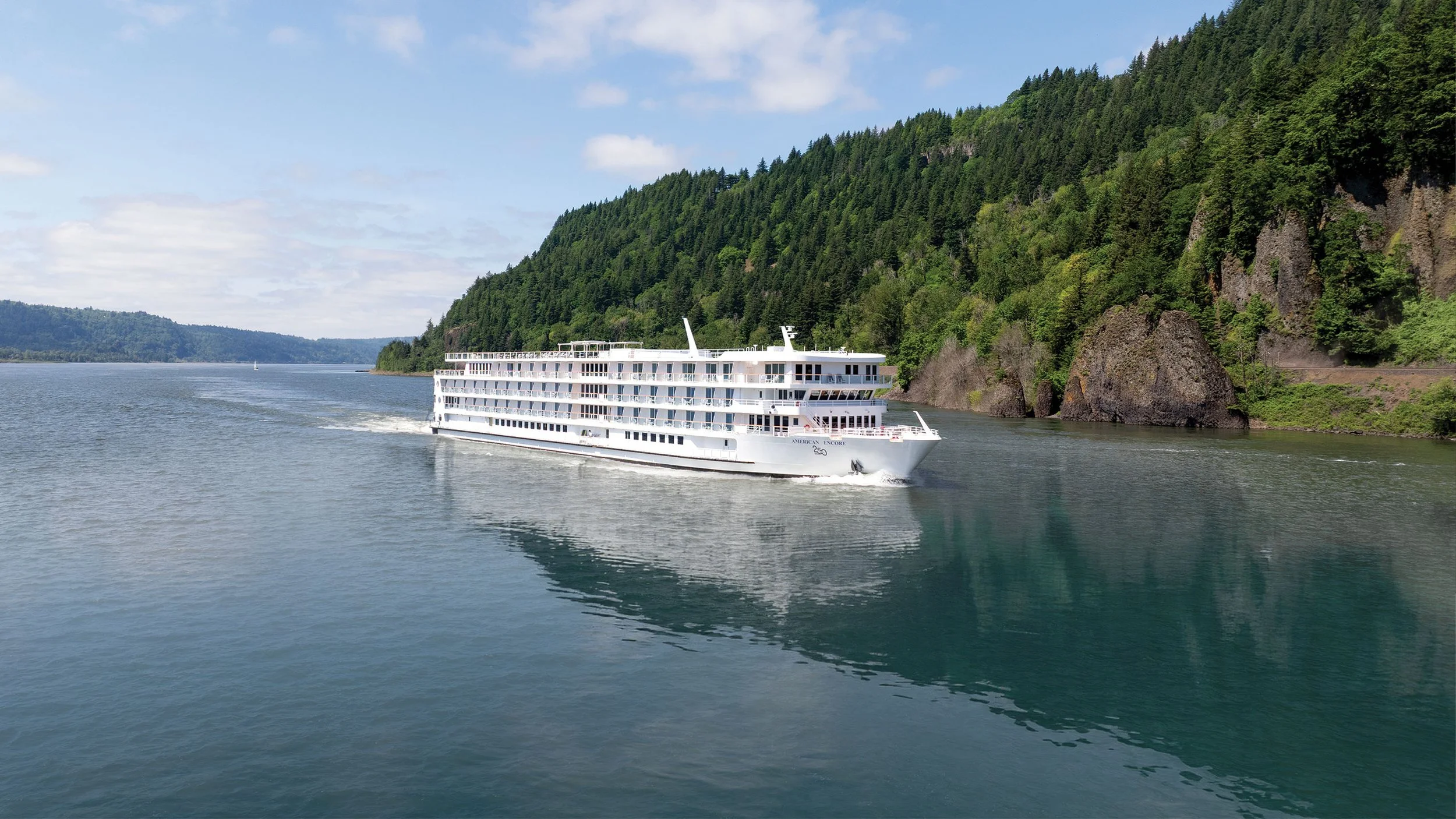 A white cruise ship sailing on a river with dense green forested hills on either side.