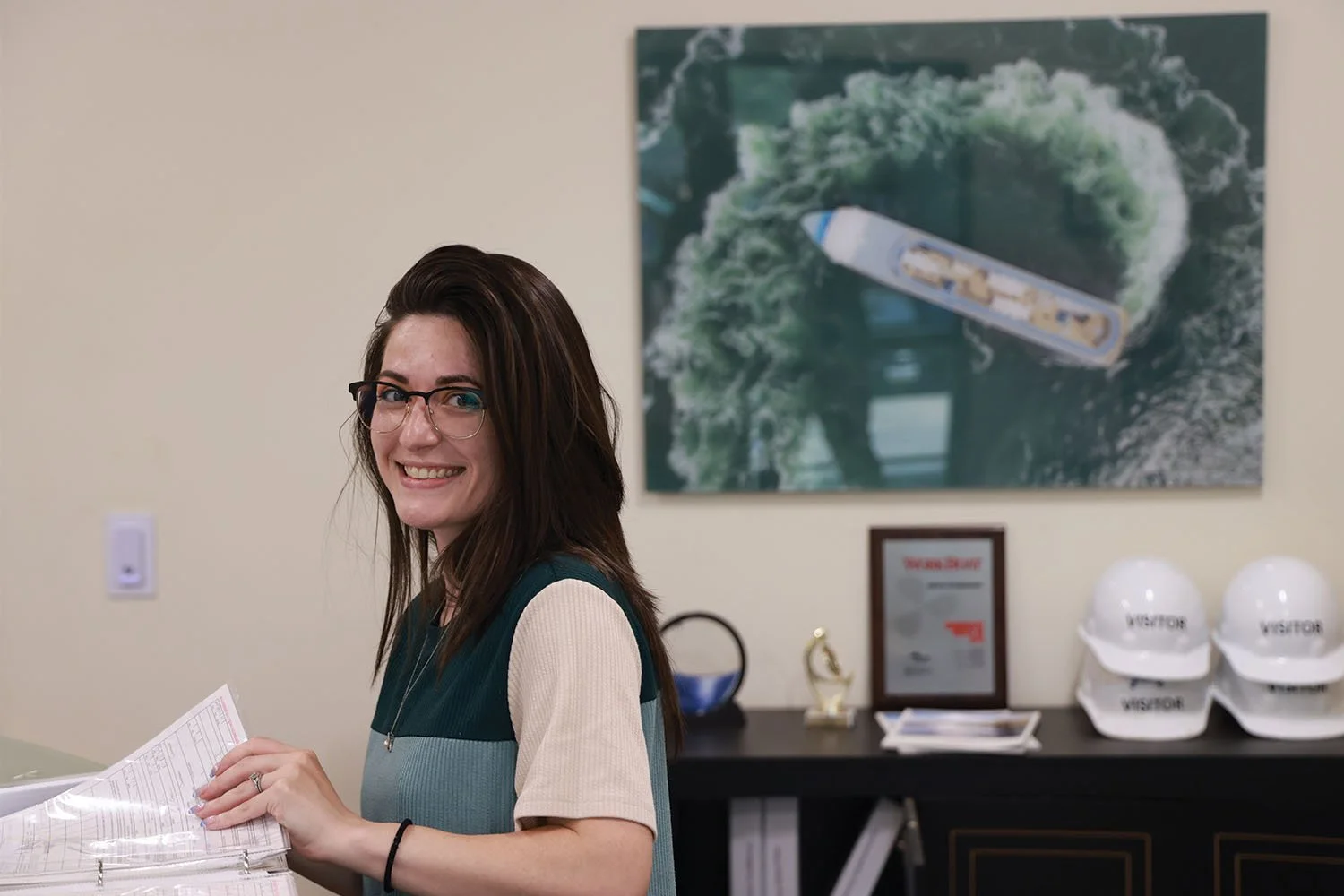 A smiling office assistant reviewing document binders at a desk with an overhead cruise ship photo behind her.