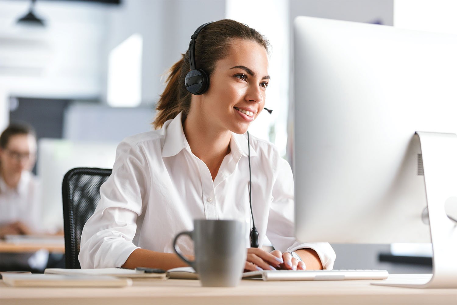 A woman wearing a headset and white shirt working at her computer in an office, smiling with a coffee mug on the desk.