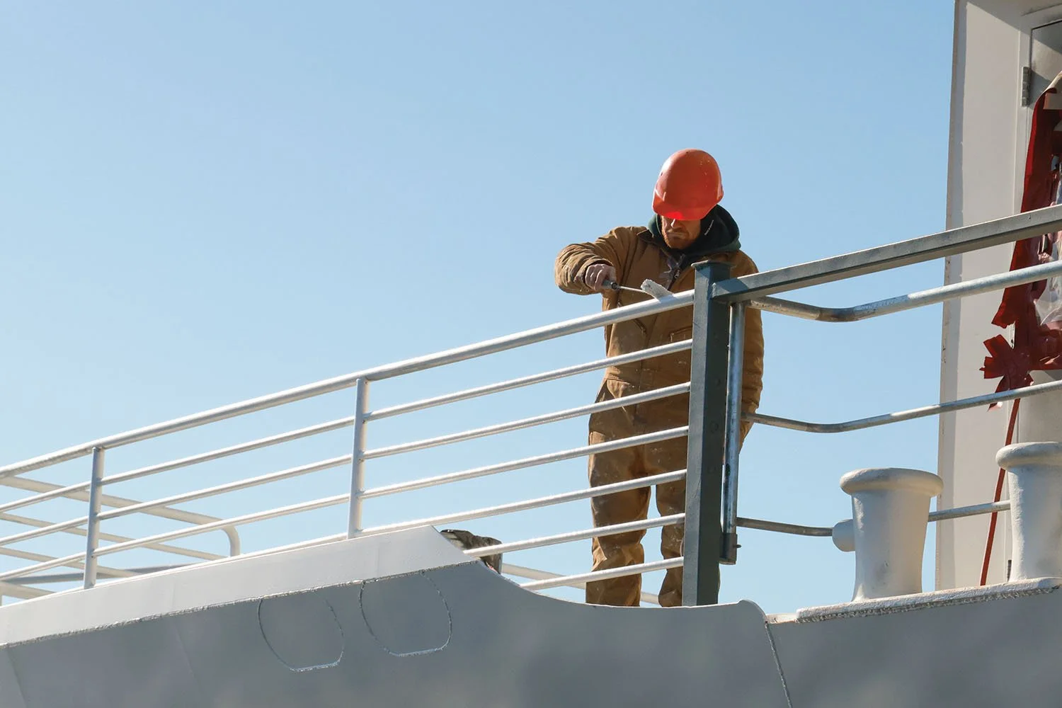 A maintenance worker in a brown jumpsuit and hard hat paints a deck railing on an upper level of a ship against a clear blue sky.