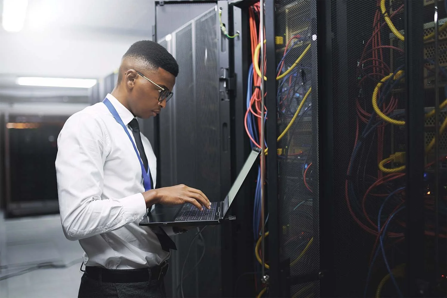 An IT professional in a white shirt and tie typing on a laptop next to server racks in a data center room.
