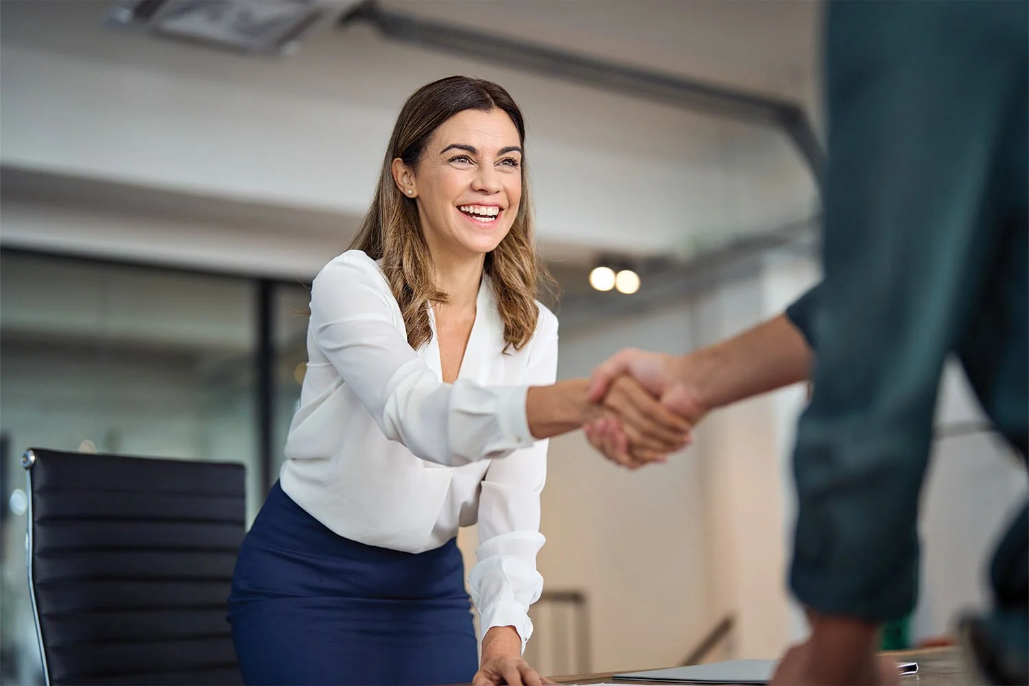 A woman with brown hair wearing a white blouse and blue skirt shaking hands with a person in a teal shirt in an office setting.