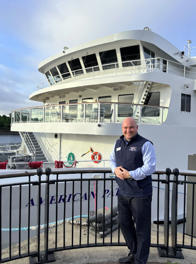 Man standing on a dock in front of an American Cruise Lines' ship named 'with multiple decks, outdoor seating, and a railing, during daytime.