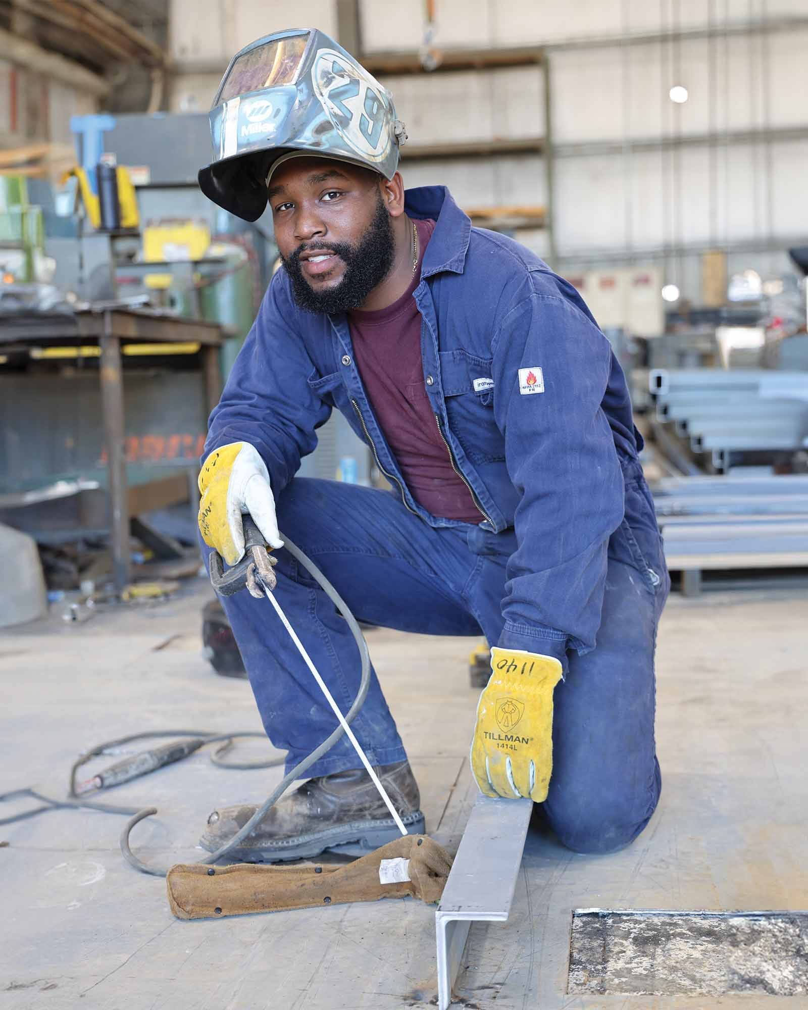A man wearing a welding helmet pushed back on his head, kneeling in a workshop. He is dressed in blue work overalls and yellow gloves. The workshop contains metal materials and tools.