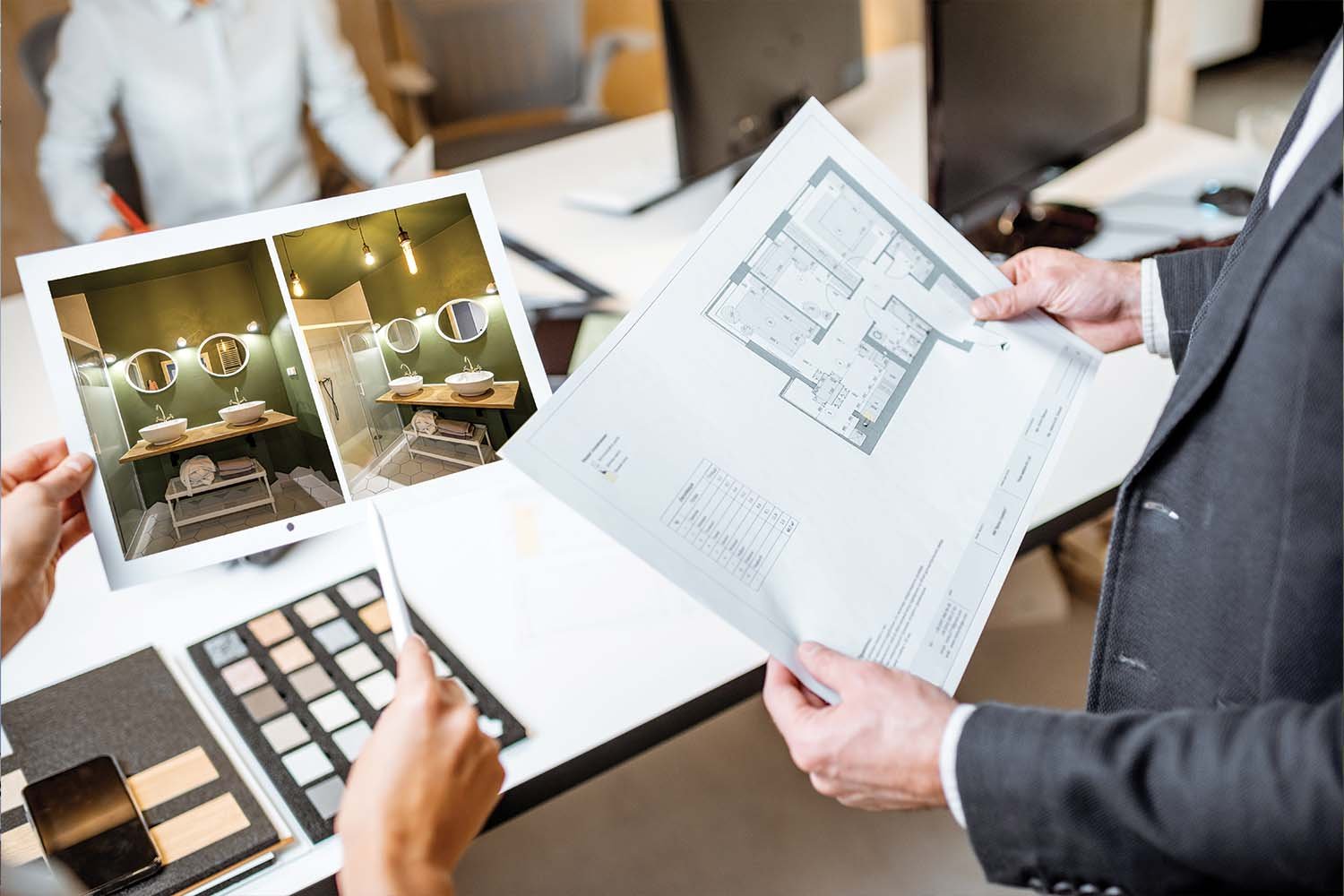 Two people discussing interior design plans, one holding a photo of a bathroom with green walls and round mirrors, the other holding architectural blueprints, with various fabric and color samples on the table.