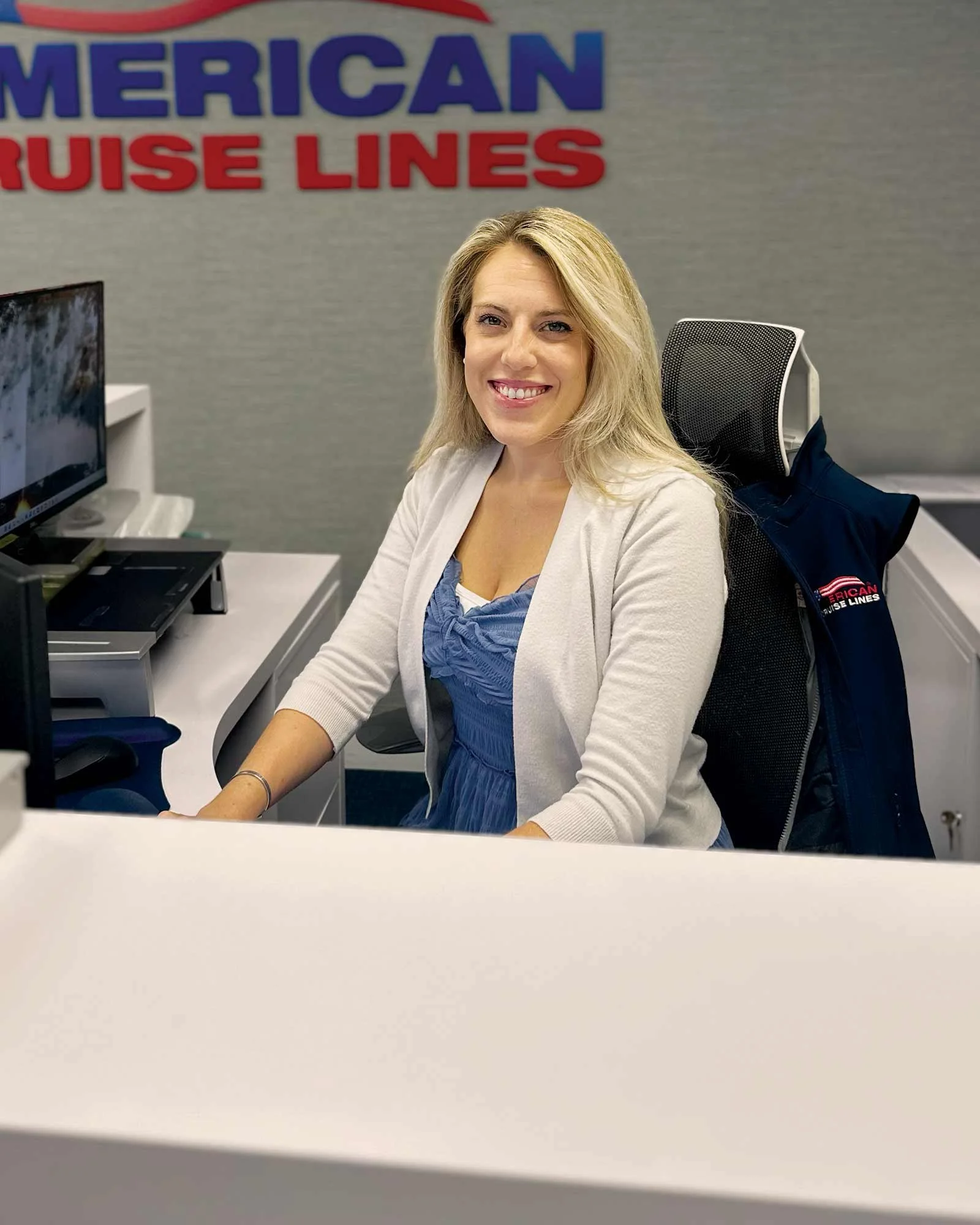 A smiling blonde woman sitting at a desk in an office with a sign that reads 'AMERICAN CRUISE LINES' on the wall behind her.