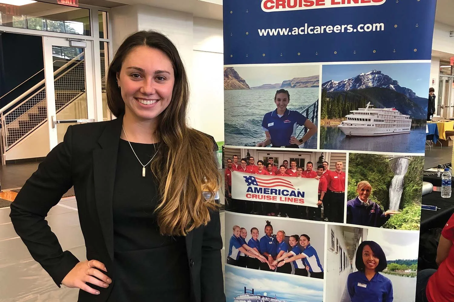 A woman with long brown hair wearing a black blazer and a necklace standing in front of a promotional display for American Cruise Lines. The display features photos of cruise ships, a group holding a banner that says "American Cruise Lines," and scenic images of lakes, mountains, and waterfalls.