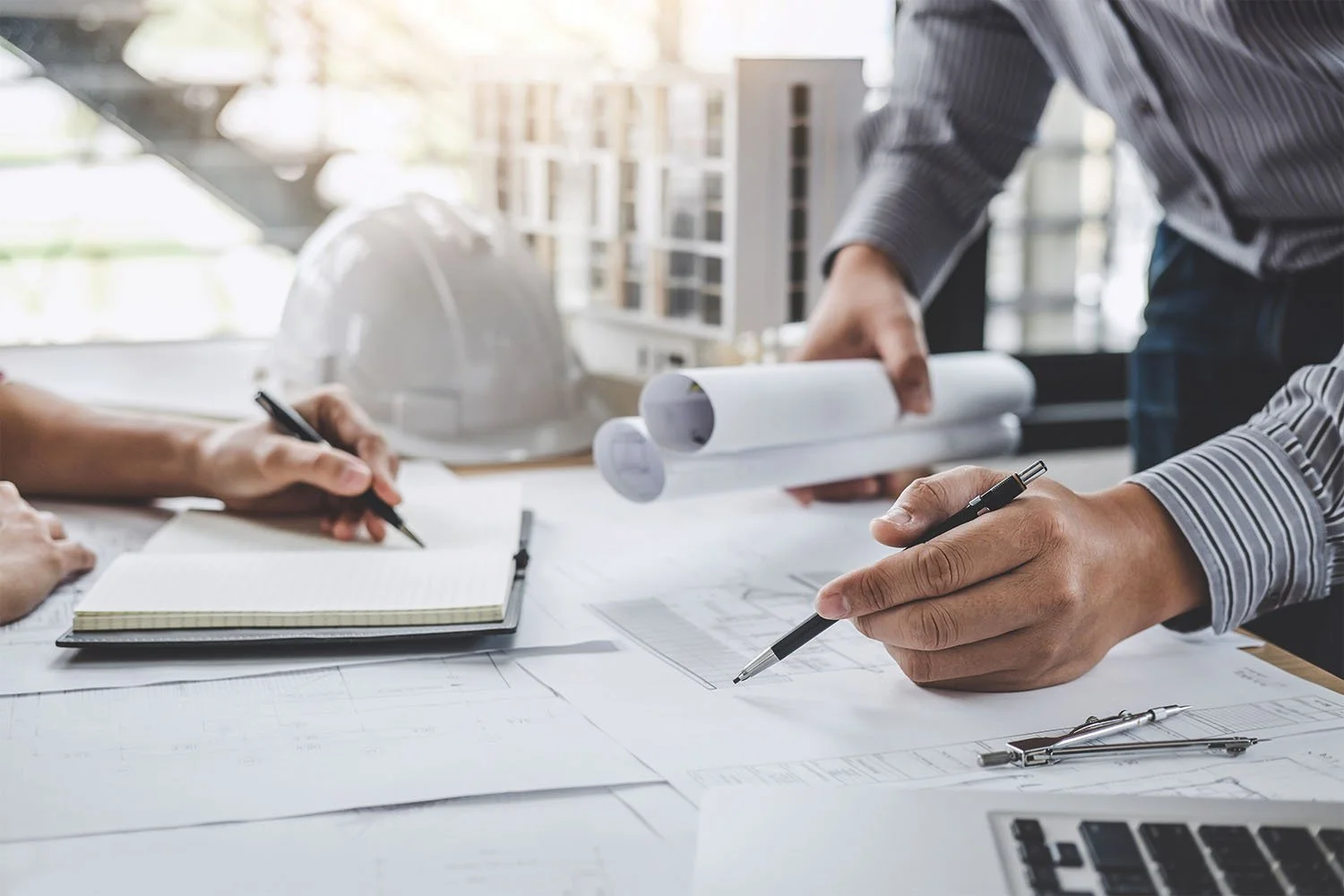 People reviewing architectural blueprints and notes at a table with a laptop, notebook, rolled blueprints, and a white hard hat in the background.