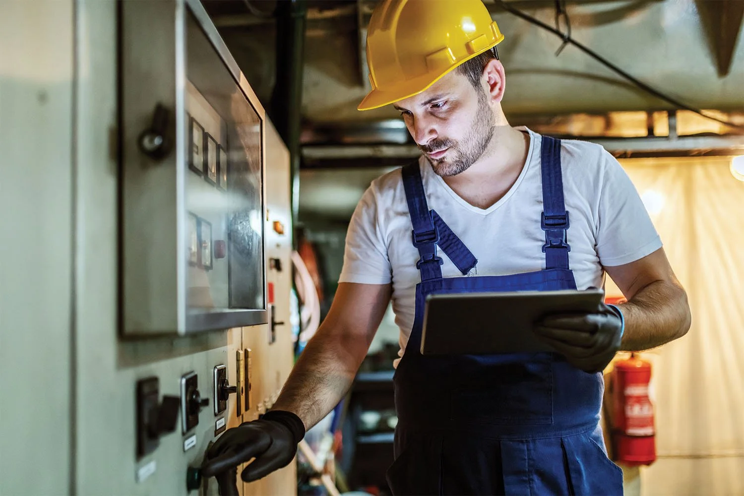 A maintenance technician wearing a hard hat and blue overalls holding a digital tablet while operating an electrical control panel.