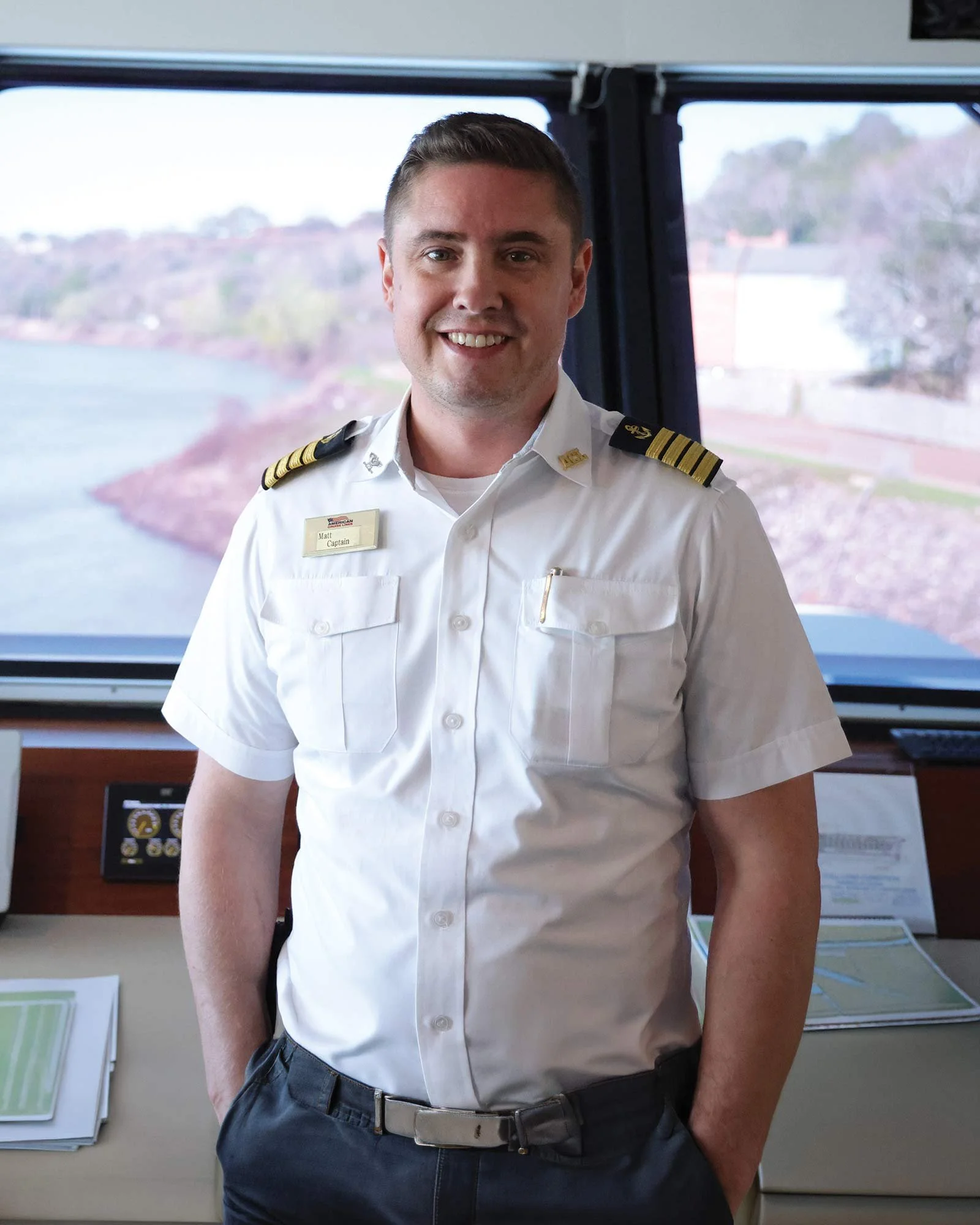 A man in a white shirt and dark pants standing indoors against a window with a scenic outdoor view, smiling, with a name tag that reads "Matt Captain".