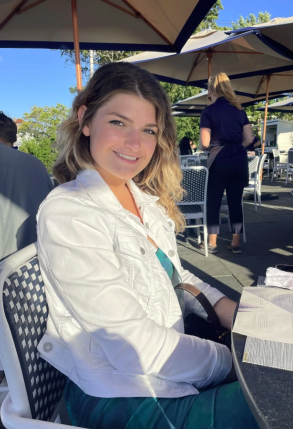 Young woman sitting at an outdoor restaurant table smiling at the camera, with sunlight casting shadows. There are large umbrellas overhead, and other patrons and staff are visible in the background.