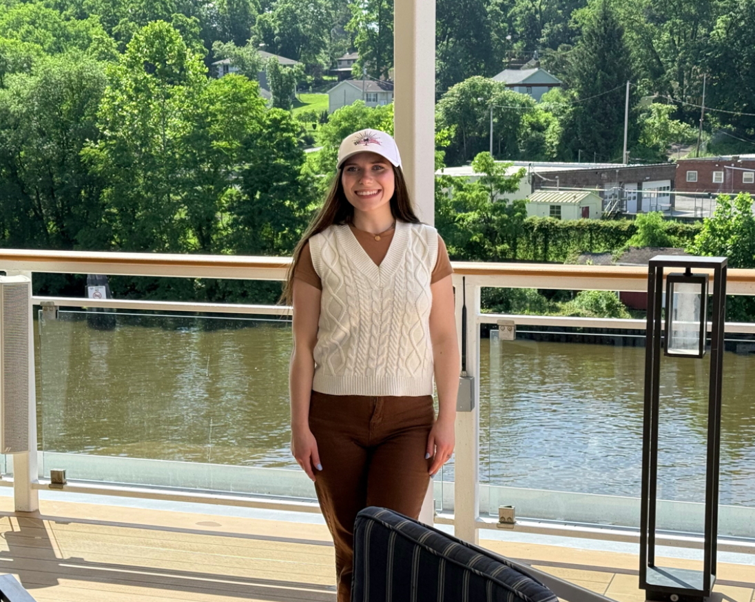 A young woman smiling, wearing a white cap, cream sweater vest over a brown shirt, and brown pants, standing on a balcony overlooking a river and lush green trees.