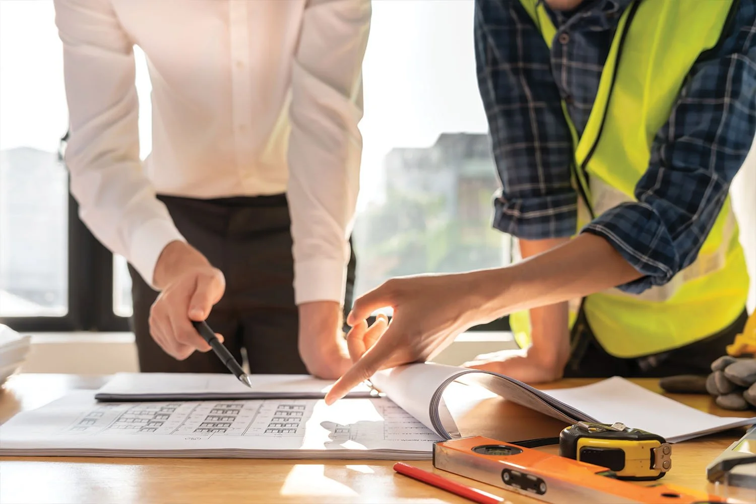 Two construction workers review blueprints at a table, one pointing at the plans with a finger and the other holding a pen, with tools and documents scattered on the table.