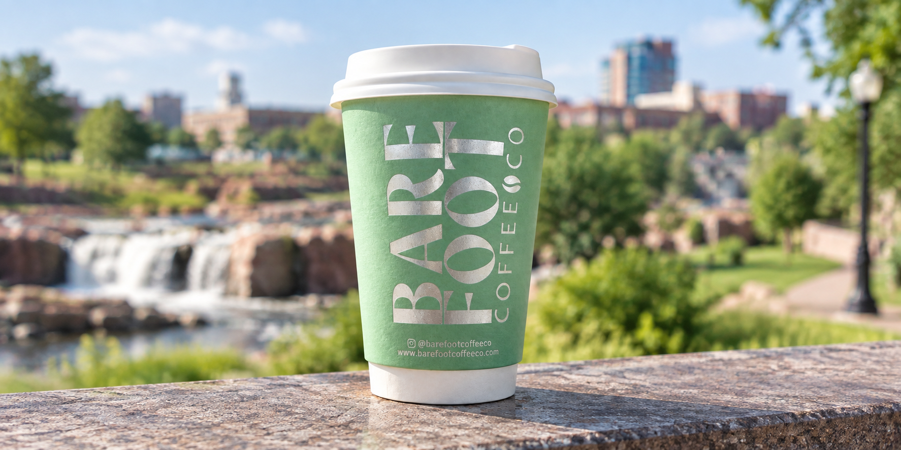 Green coffee cup with a white lid on a stone surface, with a blurred outdoor park, trees, waterfalls, and buildings in the background.
